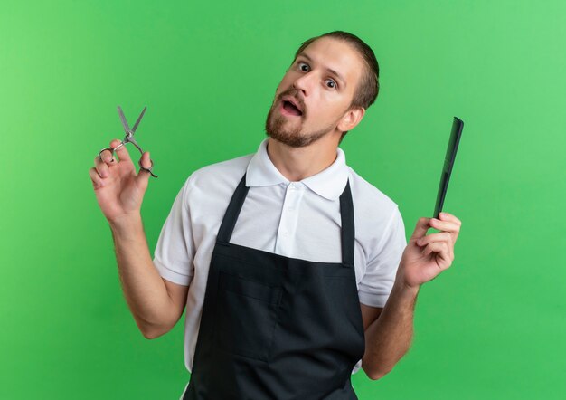 Barbero guapo joven impresionado con uniforme sosteniendo tijeras y peine aislado en verde