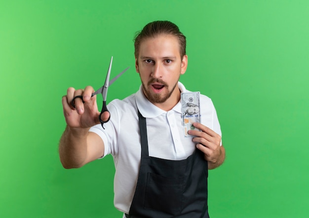 Barbero guapo joven impresionado vistiendo uniforme sosteniendo dinero y estirando tijeras aisladas en verde con espacio de copia