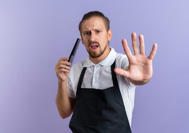 Barbero guapo joven molesto vistiendo uniforme sosteniendo el peine y gesticulando parada aislada en púrpura con espacio de copia