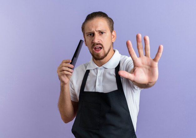 Barbero guapo joven molesto vistiendo uniforme sosteniendo el peine y gesticulando parada aislada en púrpura con espacio de copia