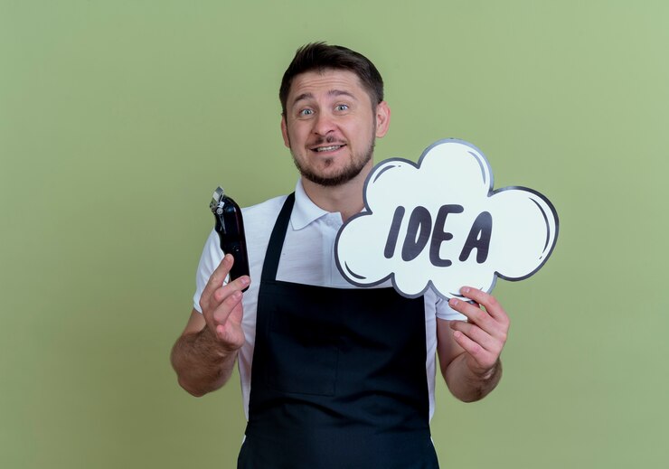Foto gratuito barbero hombre en delantal con recortador de barba y bocadillo signo palabra idea con cara feliz de pie sobre la pared verde
