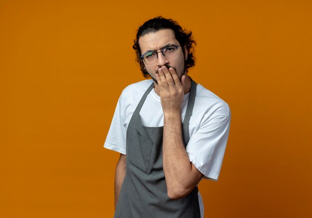 Barbero masculino caucásico joven confundido vistiendo uniforme y gafas poniendo la mano en la boca aislada sobre fondo naranja con espacio de copia