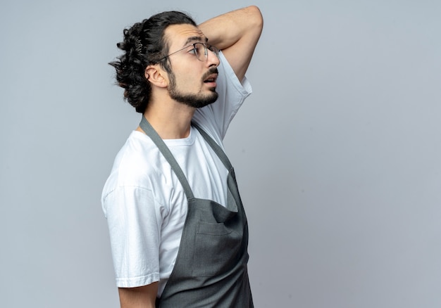 Barbero masculino caucásico joven impresionado con gafas y banda de pelo ondulado en uniforme de pie en vista de perfil mirando directamente con la mano detrás de la cabeza