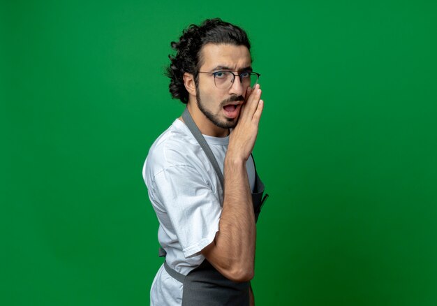 Barbero masculino caucásico joven impresionado con gafas y banda de pelo ondulado en uniforme poniendo la mano cerca de la boca susurrando a la cámara aislada sobre fondo verde con espacio de copia