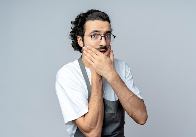 Barbero masculino caucásico joven impresionado con gafas y banda de pelo ondulado en uniforme poniendo las manos en el mentón aislado sobre fondo blanco con espacio de copia