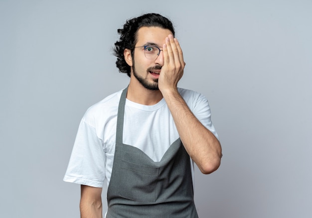 Barbero masculino caucásico joven impresionado con gafas y banda para el pelo ondulado en uniforme que cubre la mitad de la cara con la mano aislada sobre fondo blanco con espacio de copia