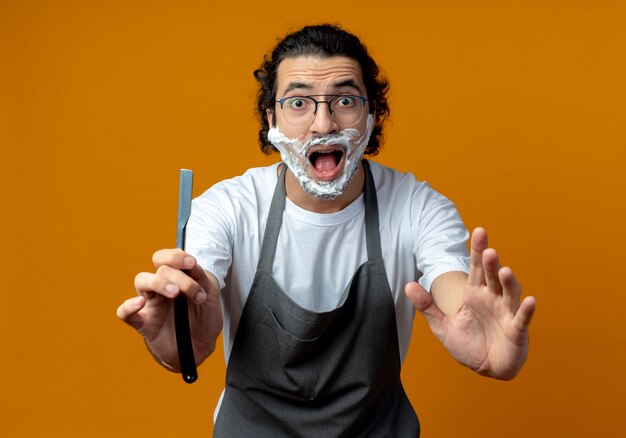 Barbero masculino caucásico joven impresionado con gafas y banda de pelo ondulado en uniforme que se extiende la navaja recta y la mano con crema de afeitar en la cara