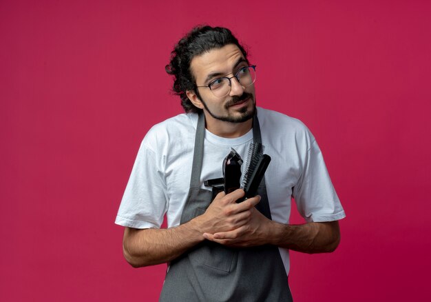 Barbero masculino caucásico joven impresionado con gafas y banda de pelo ondulado en uniforme sosteniendo herramientas de peluquero mirando al lado aislado sobre fondo carmesí con espacio de copia