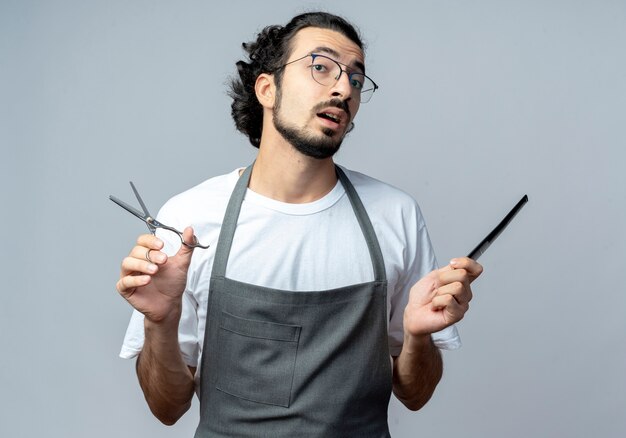 Barbero masculino caucásico joven impresionado con gafas y banda de pelo ondulado en uniforme sosteniendo tijeras y peine aislado sobre fondo blanco.