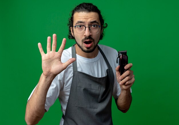 Barbero masculino caucásico joven impresionado con uniforme y gafas sosteniendo cortapelos y mostrando cinco con la mano aislada sobre fondo verde con espacio de copia