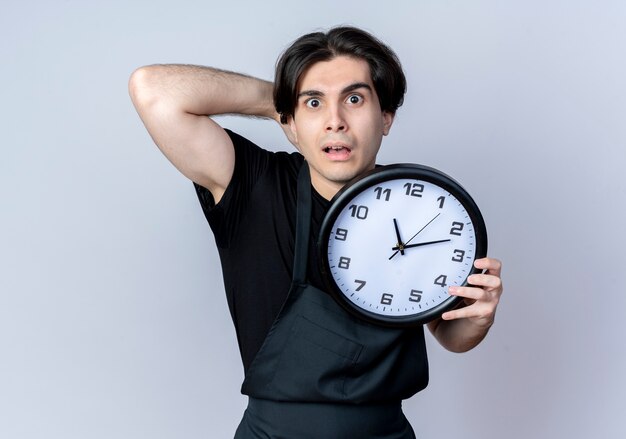 Barbero masculino guapo joven asustado en uniforme sosteniendo el reloj de pared y poniendo la mano detrás de la cabeza aislada en la pared blanca