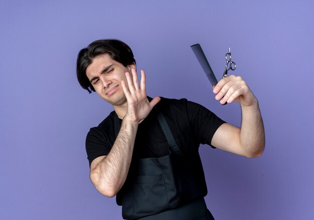 Barbero masculino guapo joven disgustado en la celebración uniforme y no mira las tijeras y el peinado aislado en azul