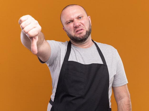 Barbero masculino de mediana edad eslavo descontento en uniforme que muestra el pulgar hacia abajo aislado en la pared naranja