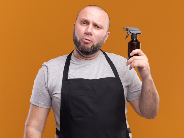 Barbero masculino de mediana edad eslavo impresionado en uniforme sosteniendo una botella de spray de agua aislada en la pared naranja