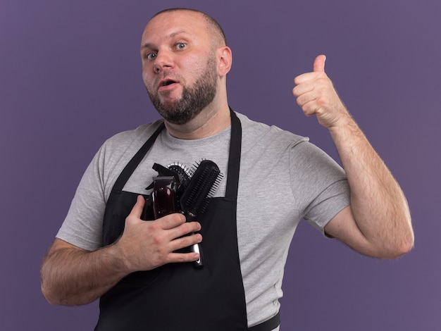 Barbero masculino de mediana edad eslavo impresionado en uniforme sosteniendo herramientas de peluquero mostrando el pulgar hacia arriba aislado en la pared púrpura