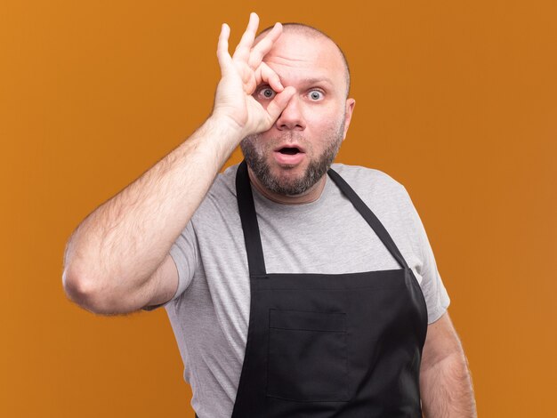 Barbero masculino de mediana edad eslavo sorprendido en uniforme que muestra el gesto de la mirada aislado en la pared naranja