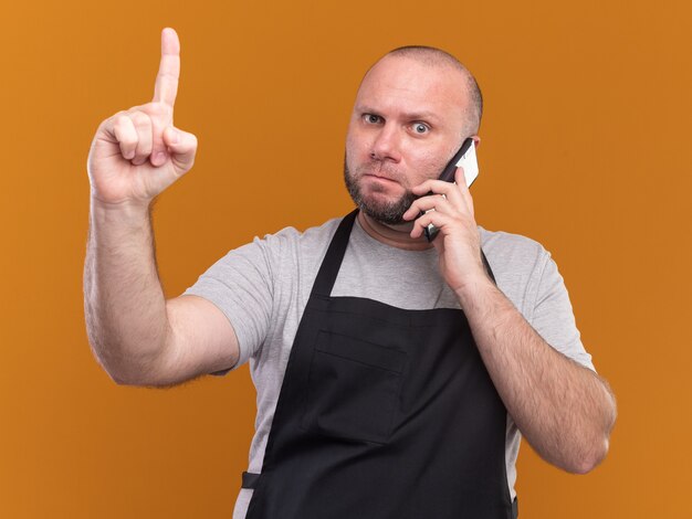 Barbero masculino de mediana edad estricto en uniforme habla en puntos de teléfono en arriba aislado en la pared naranja