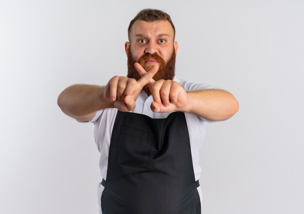 Barbero profesional barbero en delantal con cara enojada haciendo señal de stop cruzando los dedos índices de pie sobre la pared blanca