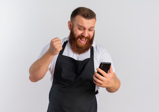 Barbero profesional barbero en delantal mirando la pantalla de su teléfono inteligente feliz y emocionado apretando el puño regocijándose de su éxito de pie sobre la pared blanca