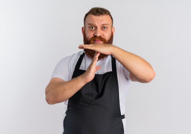 Barbero profesional barbero en delantal preocupado haciendo gesto de tiempo de espera con las manos de pie sobre la pared blanca