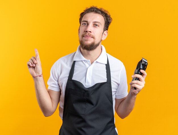 Barbero de sexo masculino joven impresionado vistiendo uniforme que sostiene los puntos de las podadoras de pelo en arriba aislados en la pared amarilla