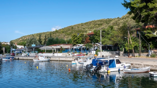 Barcos amarrados en el agua cerca de la calle del terraplén con edificios y restaurantes, mucha vegetación, colinas verdes, Grecia