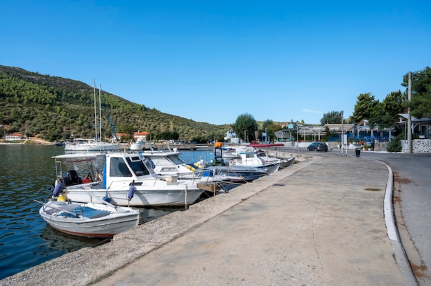 Barcos amarrados en el agua cerca de la calle del terraplén con edificios y restaurantes, mucha vegetación, colinas verdes, Grecia