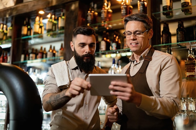 Barista adulto medio y su joven compañero de trabajo usando el panel táctil mientras trabajan en un pub