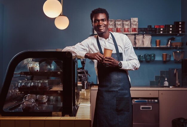 Barista africano sonriendo a la cámara relajándose después del día de trabajo con café mientras se apoya en el mostrador de la cafetería.