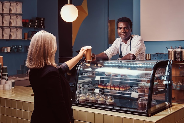 Barista afroamericano uniformado dando una taza de café a su cliente en la cafetería de moda