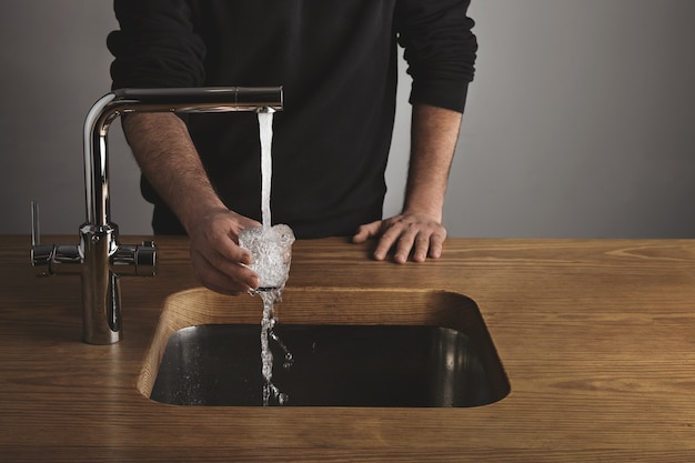 Barista brutal en sweatshot negro detrás de una mesa de madera gruesa enjuaga un pequeño vaso transparente con agua bajo el grifo de metal plateado en la cafetería. Gotas de agua de un vaso.