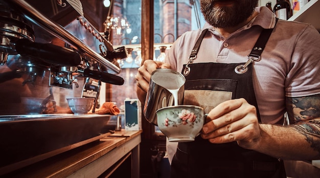 Barista en delantal haciendo un capuchino, sirviendo leche en una taza en un restaurante o cafetería