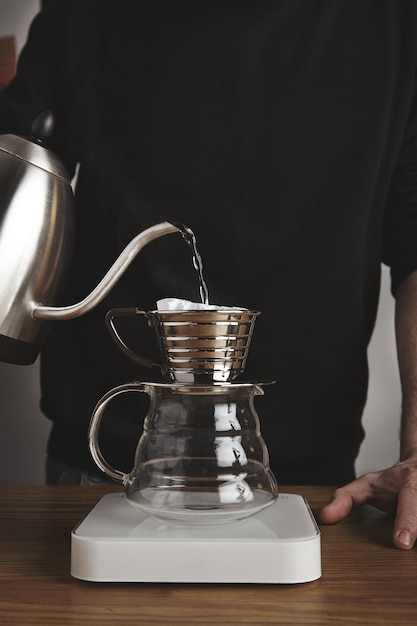 barista derrama agua caliente para preparar café filtrado desde una tetera moderna hasta una hermosa cafetera de goteo transparente sobre pesas blancas simples.