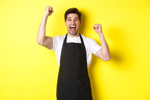 Barista feliz celebrando la victoria, levantando las manos y gritando de alegría, vestido con delantal negro, uniforme de tienda, de pie contra el fondo amarillo.