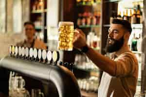 Foto gratuita barista feliz sosteniendo un vaso de cerveza de barril mientras trabaja en el mostrador del bar