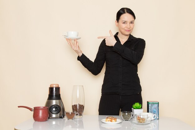 Barista femenina en pantalón negro delante de la mesa con equipo de ingredientes de café en la pared blanca