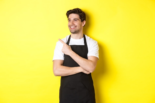 Barista guapo apuntando y mirando a la izquierda en la promoción, vestido con delantal negro, de pie contra el fondo amarillo