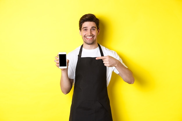 Barista guapo en delantal negro apuntando con el dedo a la pantalla del móvil, mostrando la aplicación y sonriendo, de pie sobre fondo amarillo.
