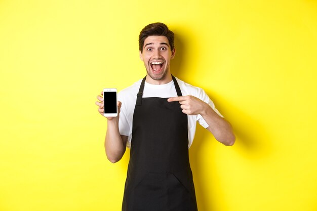Barista guapo en delantal negro apuntando con el dedo a la pantalla del móvil, mostrando la aplicación y sonriendo, de pie sobre fondo amarillo.