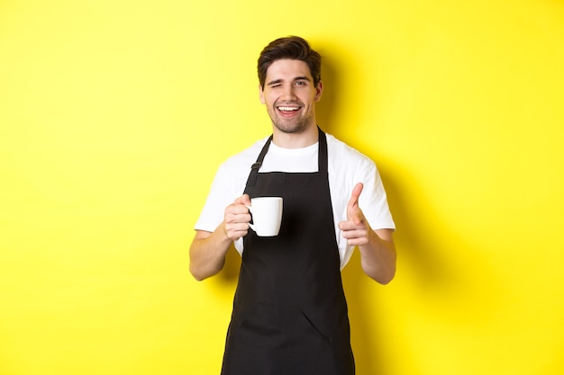 Barista guapo en delantal negro sosteniendo la taza de café, señalando con el dedo, invitando a visitar café, de pie sobre fondo amarillo