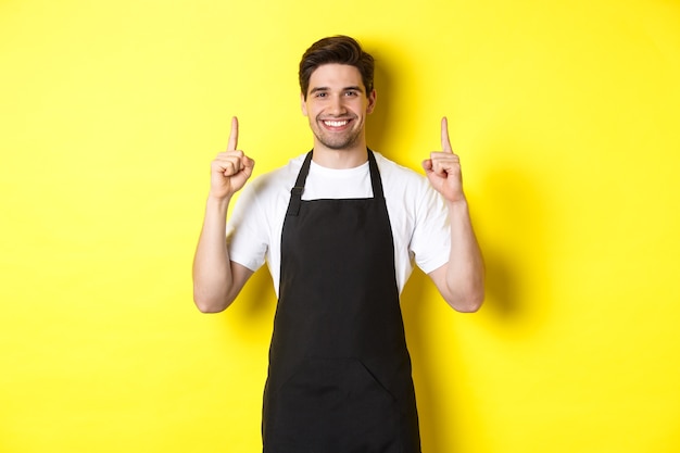 Barista guapo mostrando publicidad, oferta promocional de cafetería, apuntando con el dedo hacia arriba, de pie contra el fondo amarillo.