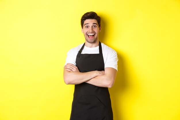 Barista guapo sorprendido en delantal negro levantando las cejas, mirando asombrado, de pie contra el fondo amarillo.