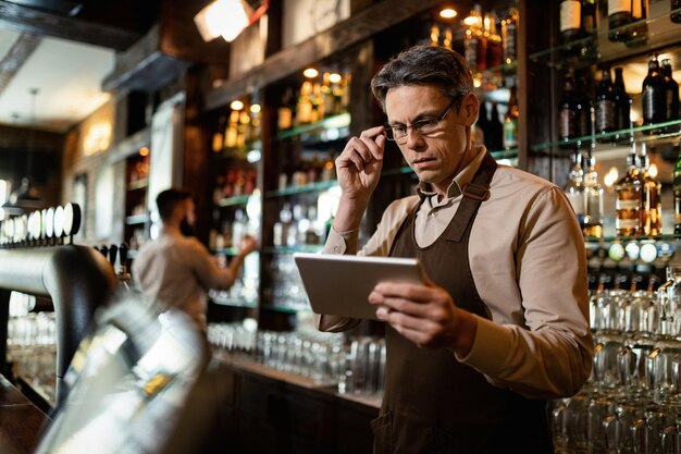 Barista de mediana edad que usa una tableta digital mientras trabaja en un bar. Su compañero de trabajo está en segundo plano.