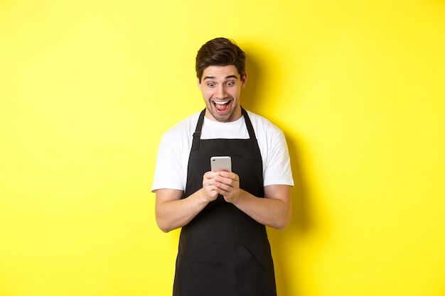 Barista mirando sorprendido mientras lee el mensaje en el teléfono móvil de pie en delantal negro contra amarillo ...