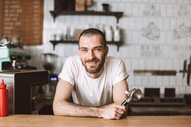 Barista sonriente apoyado en el mostrador del bar y felizmente mirando a la cámara mientras sostiene el portafiltro en la mano en el café