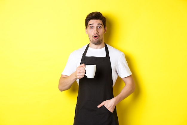 Barista sosteniendo la taza de café y mirando sorprendido, de pie en uniforme de café delantal negro sobre fondo amarillo.