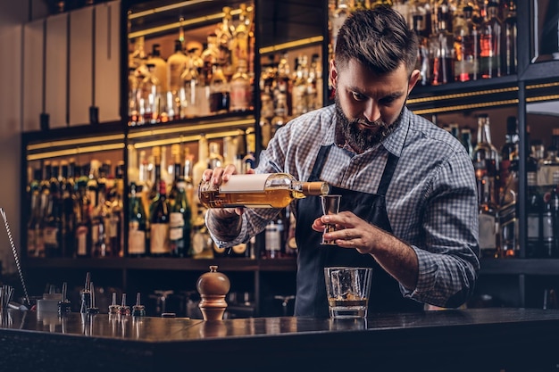 El barman brutal y elegante con una camisa y un delantal hace un cóctel en el fondo del mostrador del bar.