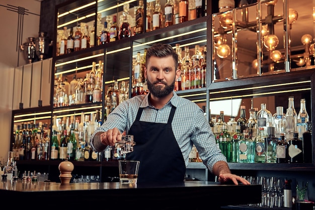 El barman brutal y elegante con una camisa y un delantal hace un cóctel en el fondo del mostrador del bar.