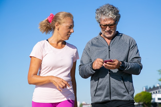 Foto gratuita basculador masculino enfocado usando la aplicación de fitness en el teléfono celular después de trotar. pareja madura vistiendo ropa deportiva, de pie afuera. comunicación y gadget para el concepto de deporte.