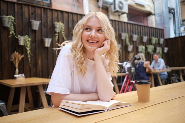 Bastante alegre chica rubia casual felizmente mirando hacia otro lado con un libro y café en la mesa en el patio de la cafetería